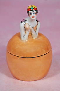 PORCELAIN ART D?CO FIGURIAL POWDER JAR BY NORITAKE: PORCELAIN ART DÉCO FIGURIAL POWDER JAR BY NORITAKE. 6”. Marks: Noritake trademark symbol. Porcelain lusterware figural powder jar features flapper lady with sculpted black hair, and