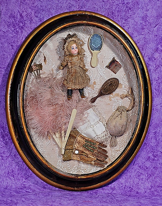 MINIATURE BISQUE DOLL WITH ACCESSORIES IN FRAMED DISPLAY.: MINIATURE BISQUE DOLL WITH ACCESSORIES IN FRAMED DISPLAY. 17” frame, 5 ½” doll. Oval shadow box display featuring German bisque socket head doll with blue glass eyes, painted lashes,