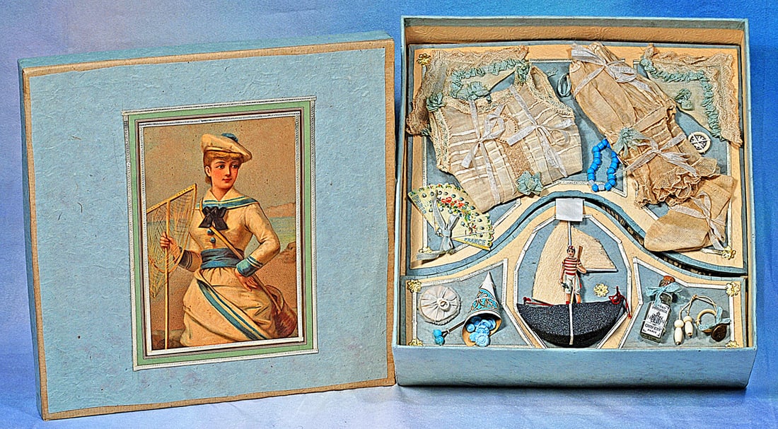 DOLL TROUSSEAU IN PRESENTATION BOX: 10 ½” x 10 ½” x 4”. Paper covered heavy cardboard box with lithographed image on box lid; opens to reveal assorted toiletries, accessory items, and under garments, tw