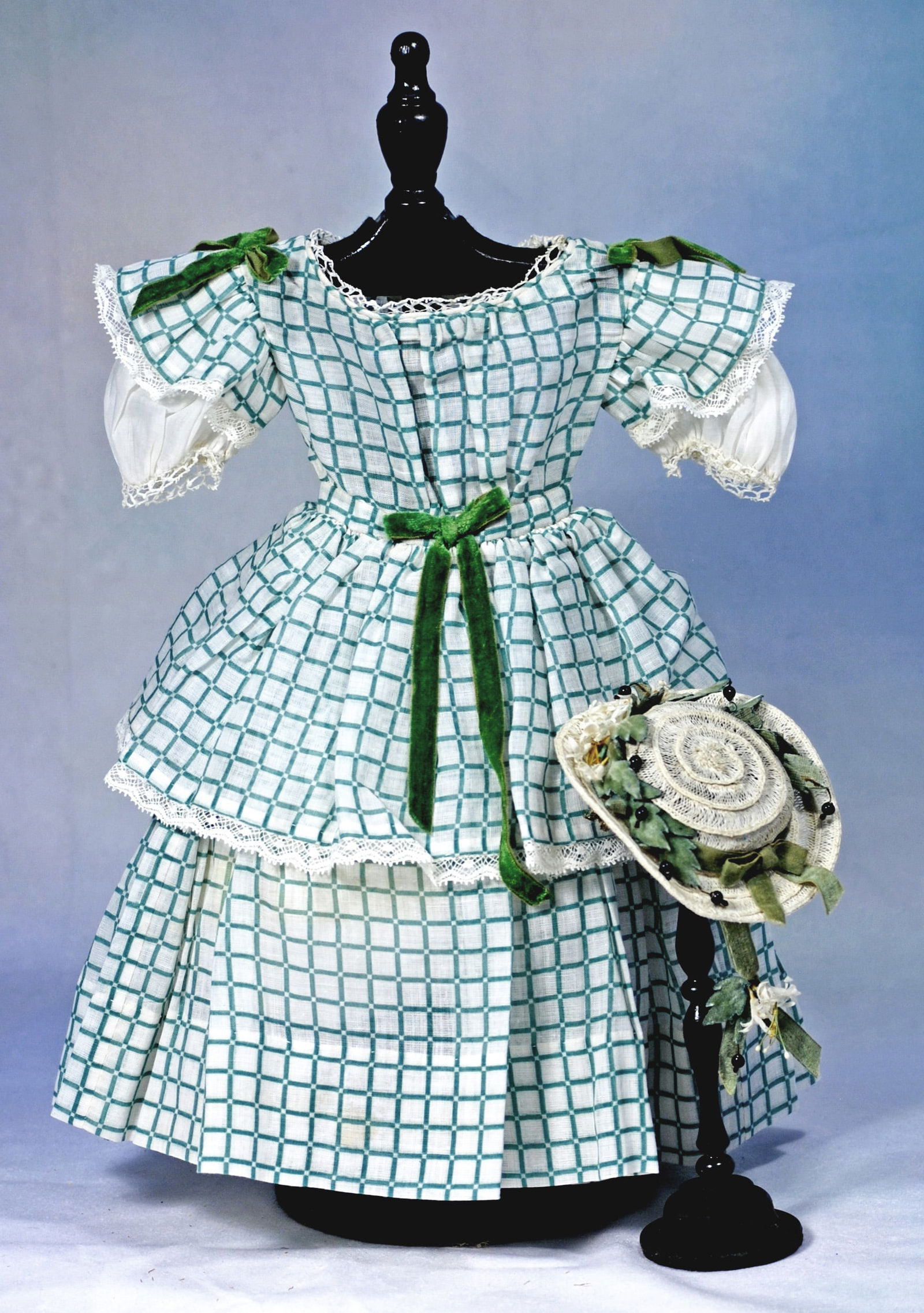 FINE GREEN AND WHITE DOLL DRESS WITH BONNET: Dress of green and white window pane fabric has rounded neckline, inset waistband, flared sleeves with lace edging over white cotton puffed sleeves, gathered full skirt with short gathered overskirt,