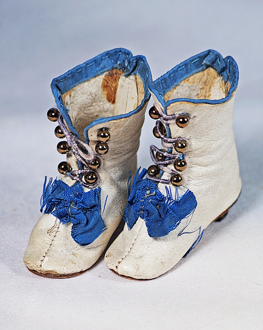 PAIR OF HURET-ERA WHITE KIDSKIN BOOTS: 2 ¼” length, White kidskin boots with teal blue silk edging and bows, 8 brass button front closing, tiny wooden heels, marked “4”.