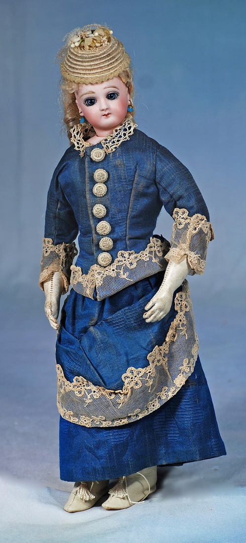 PETITE FRENCH BISQUE POUPEE IN ORIGINAL GOWN: 12 ½”. Marks: None visible. Bisque swivel head on bisque shoulderplate, large blue paperweight eyes, painted lashes, eyeliner, feathered brows, closed mouth in pert smile, pierced