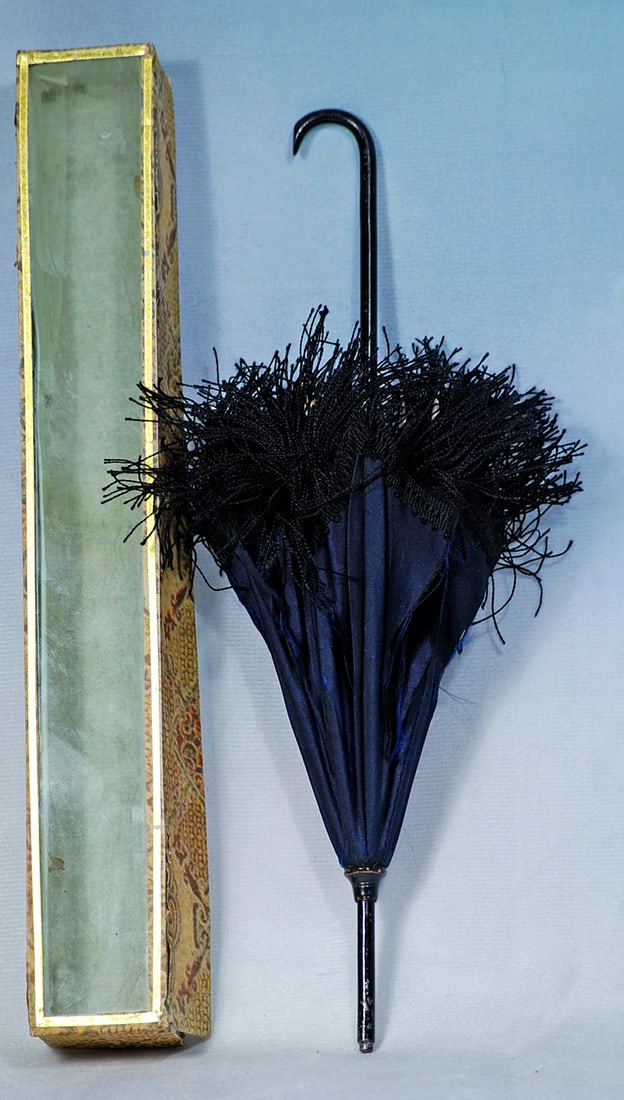 NAVY SILK PARASOL IN ORIGINAL BOX: 9 ½”. Dark navy blue silk parasol has black enamel metal handle and tip, long thick black fringe, bone tips and metal fittings, original gilt-edged cardboard box with glass front.