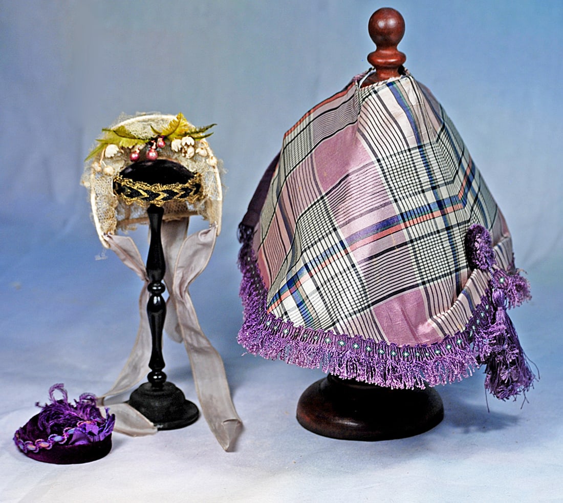 SILK TAFFETA CAPELET AND TWO DOLL BONNETS: Lavender silk plaid taffeta capelet, lined in lavender silk and enhanced with generous fringe of deep purple, and fancy round cluster with tassels decorating the back. 6 ½” length; white