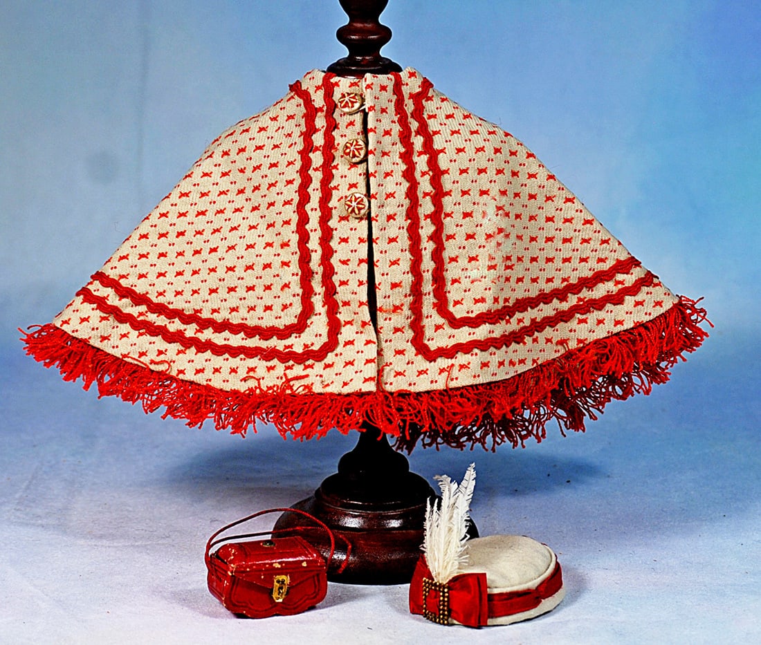 CREAM AND RED PATTERNED WOOL CAPE FOR POUPEE:  Cream and red wool made up in an attractive cape trimmed with double rows of red wool braid all around, edged in red wool fringe, with red roundelle and tassels at the back, three buttons in