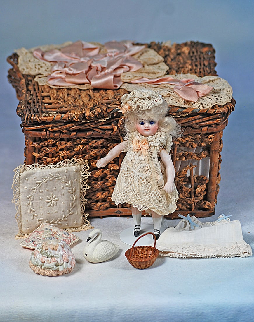 TINY BISQUE MIGNONETTE WITH WICKER BASKET: Woven wicker basket with hinged cover, padded and shirred pink silk interior, contains 4 ½” all-bisque doll with swivel neck, five-piece slender jointed bisque body, painted white