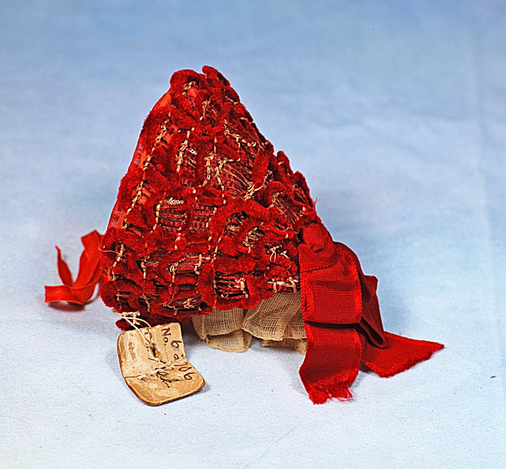 ANTIQUE RED SILK AND VELVET DOLL BONNET: Wire-framed and cone-shaped bonnet has stripes of red silk over tan base and further decorated with intricately arranged ribbons of deep red velvet with scalloped detail framing the front, red silk