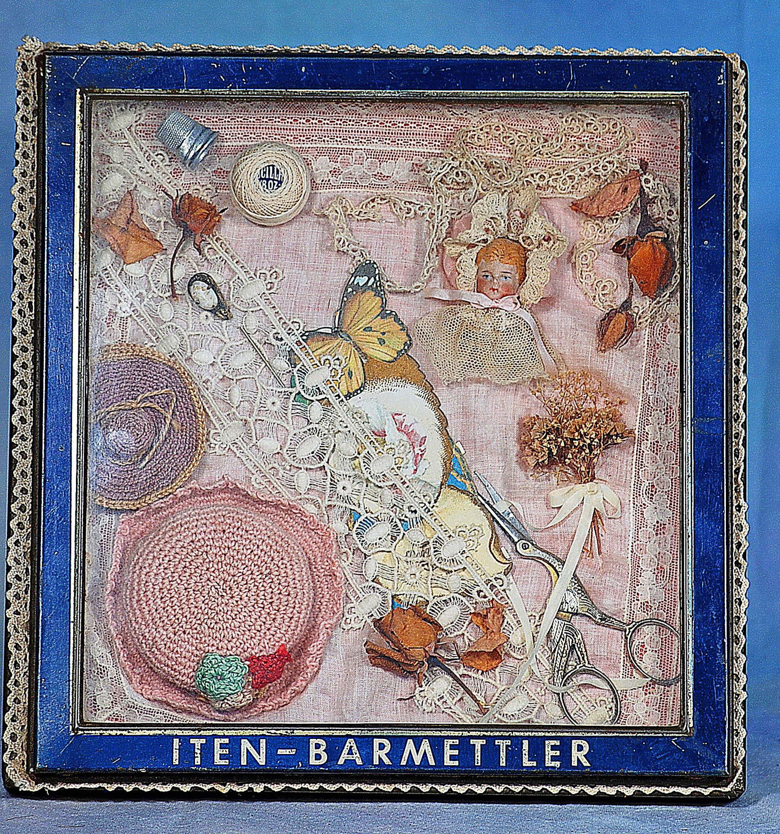 PRESENTATION DISPLAY OF MINIATURE SEWING TOOLS AND RELATED ITEMS: Glass front display case which is hinged at the side, labeled “Iten-Barmettler”, 10 ½” x 10 ½”. Display board with silk covering featuring assorted sewing and needlew