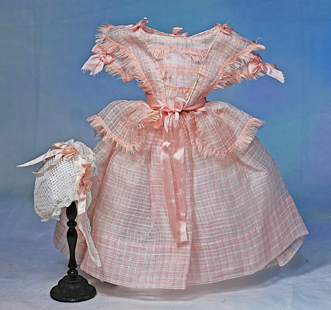 A PINK AND WHITE COTTON GOWN FOR POUPEE WITH HAND-TIED NET BONNET: Sheer muslin window-pain plaid gown in mode enfantine-style has graceful fringe-trimmed bretteles and full basque, soft pink ribbons and bows set all around, plus silk streamers, sized for