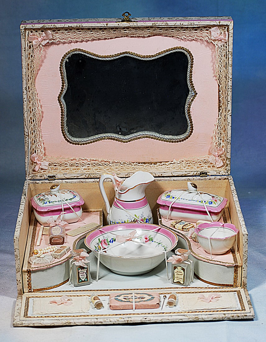 FRENCH PORCELAIN TOILETTE SET IN PRESENTATION BOX: 11 ½” x 7 ½” x 4 ½”. A wooden box covered with beautiful lithographed papers hinges open to reveals an interior with lace-edged mirror lid, 7-piece porcelain