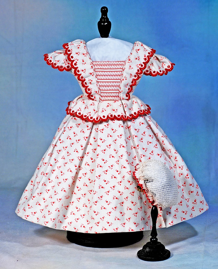 STYLISH RED AND WHITE COTTON GOWN AND HAND-TIED NET BONNET: Fine white cotton imprinted with dainty red flowers made up in a gown having plastron of striped fabric, bretelles and basque around the waist, all accented with red scalloped edging, gathered full sk
