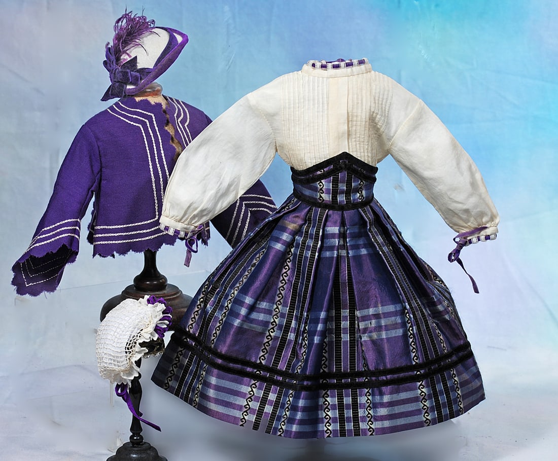 RICH PURPLE SILK AND WOOL ENSEMBLE FOR POUPEE: Features a skirt of purple and black crossed with bars of blue, high pointed waistband, pleated skirt and narrow black velvet trims; white cotton blouse with purple ribbon trim and a purple short wool