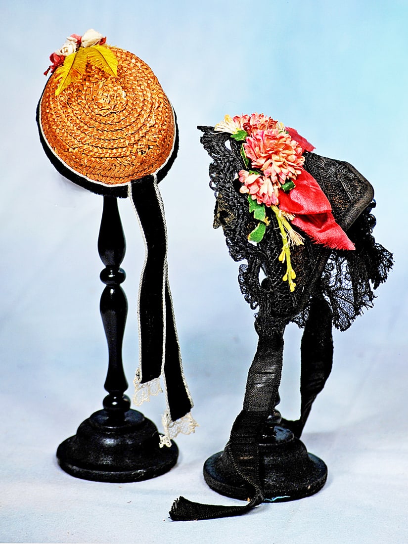 TWO DOLL BONNETS: Black straw bonnet with scalloped black lace edging, adorned with pink flowers and greenery, white lace framing the inside with black lace bavolet and silk ribbon ties; plus a woven natural straw