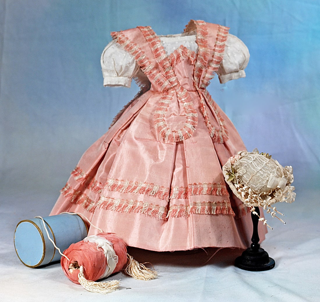PINK SILK ENSEMBLE FOR POUPEE: Features white dotted Swiss gown with rounded neckline and shor puffed sleeves under a soft pink silk taffeta gown with bretelles set over a pointed high waistband and extending over the back falling