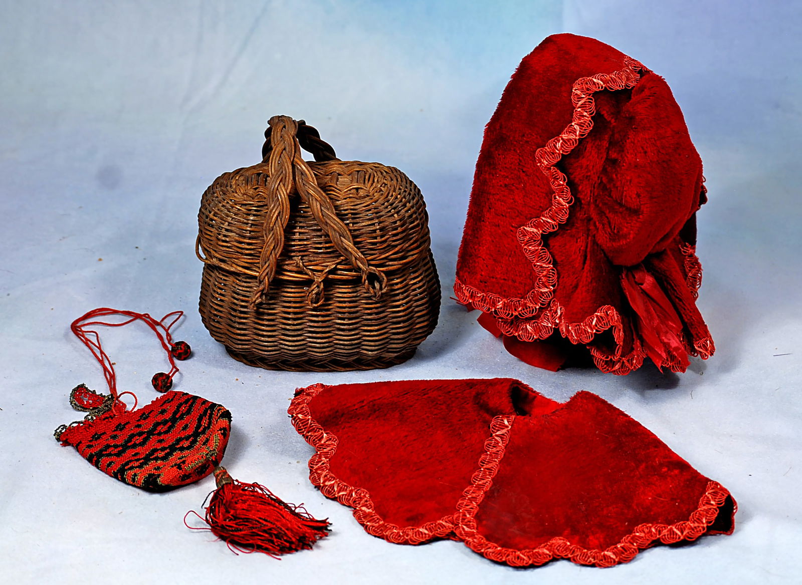 ACCESSORIES FOR FRENCH POUPEE OR BEBE: Including a 4” woven wicker market basket with twin handles and hinged top; a deep red velvet hooded bonnet with silk scalloped edge trim and red silk bow and ribbons, matching cape; red and bla