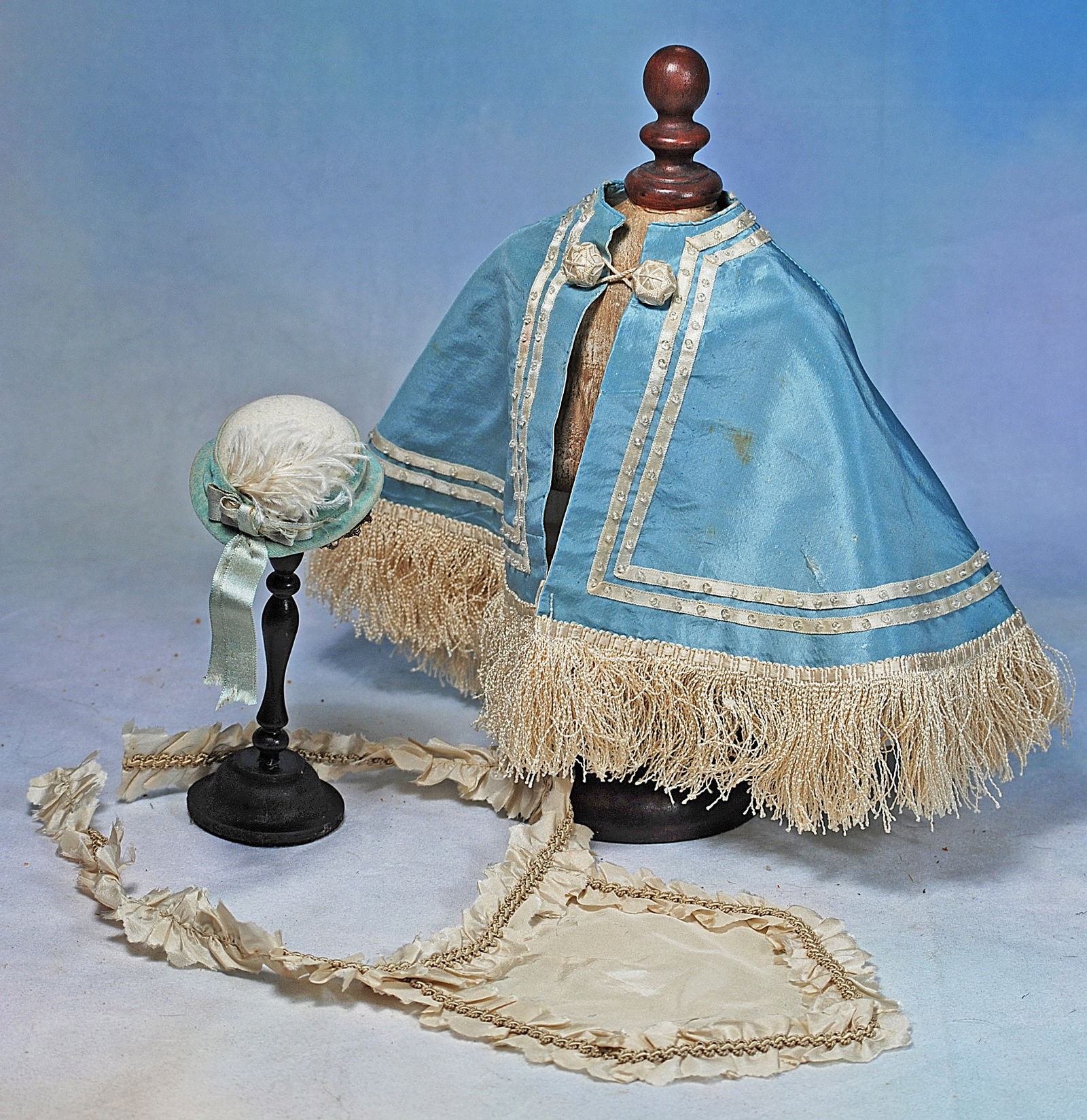 SILK CAPE, HAT, AND SATIN APRON FOR POUPEE: Cream silk taffeta apron with ornate braid accenting tightly gathered ruffles and ruffled waistband, crinkled white fringe trim all around, silk ribbons at center back; white felt hat with rolled