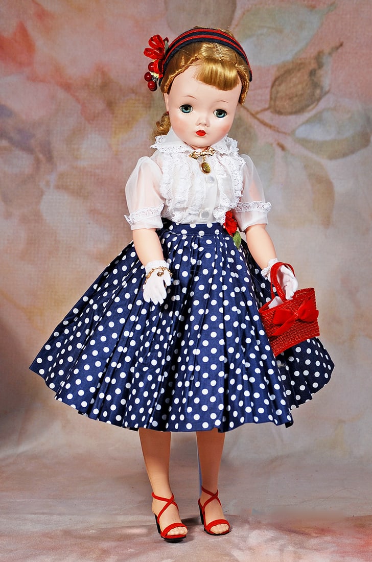 ALEXANDER CISSY IN POLKA DOT SKIRT AND LACE BLOUSE: 20”. Hard plastic socket head, adult lady body, sleep eyes, applied and painted lashes, ruby lips, blonde hair with bangs and hair pulled to the back secured by red ribbon, wearing navy blue and