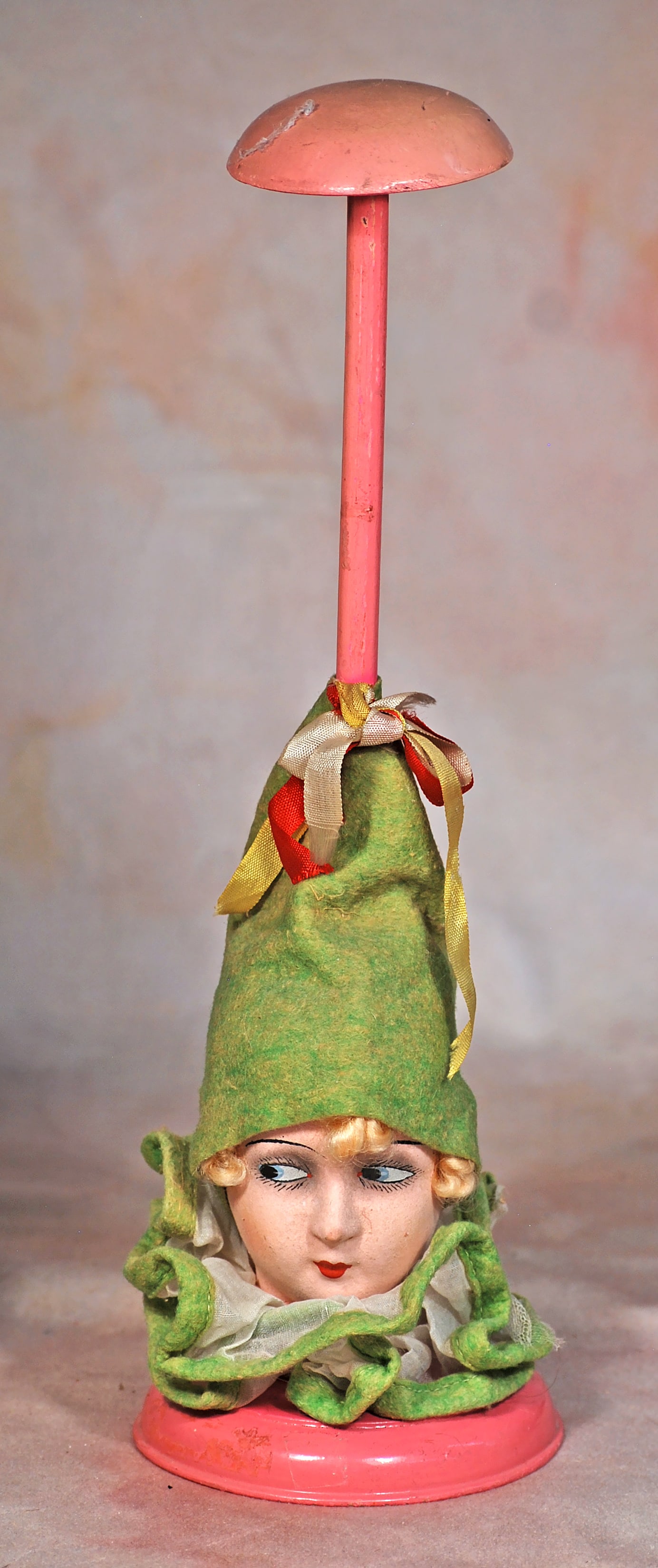 VINTAGE HAT STAND IN THE ART DECO MANNER.: 67. VINTAGE HAT STAND IN THE ART DECO MANNER. 13 ½”. Wooden hat stand with painted pink finish and featuring a paper mache doll head with painted features, side-glancing eyes, eye