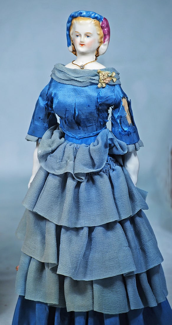 GERMAN BISQUE LADY WITH BLUE AND MAGENTA DECORATED SNOOD: 13”. Porcelain shoulderhead with sculpted blonde hair waved from face and captured under a defined sculpted snood with magenta feather and blue draped ribbon with white tips, painted blue eyes, 