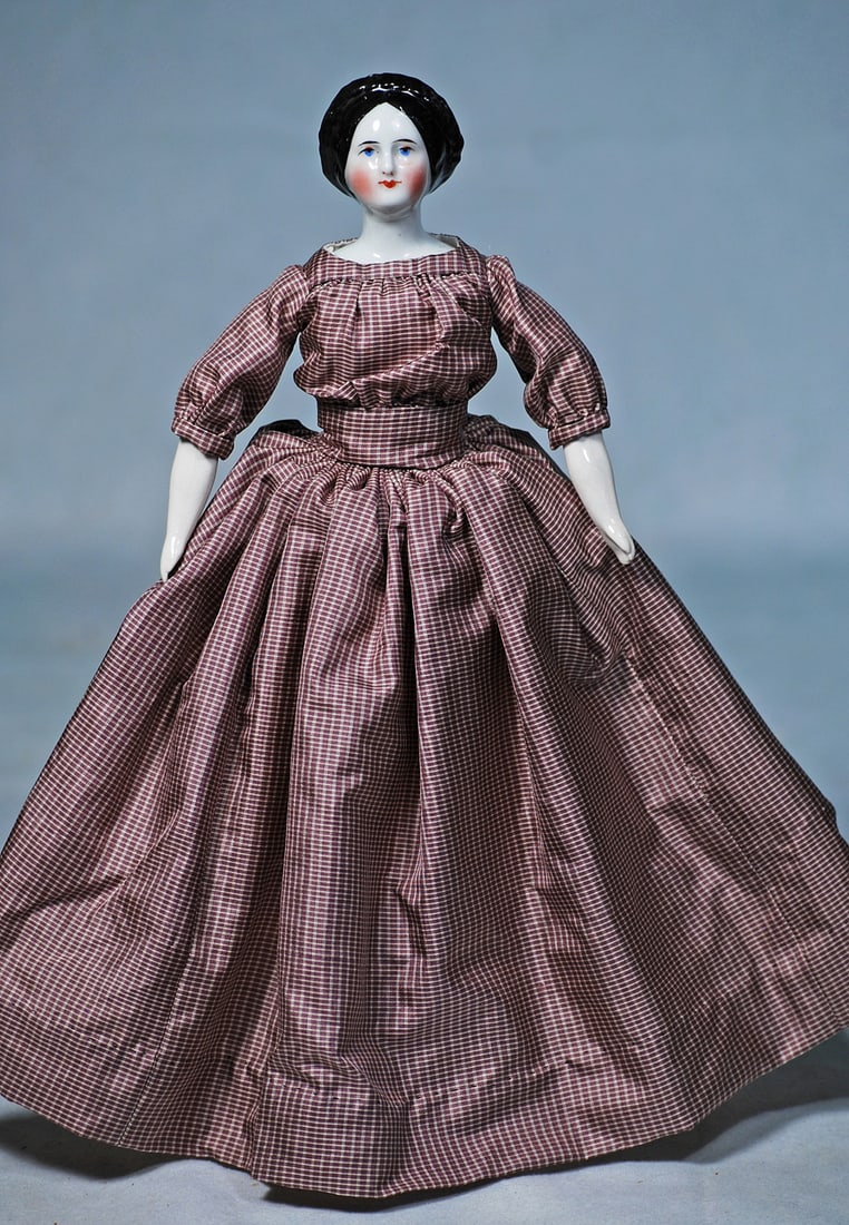 PETITE GERMAN PORCELAIN DOLL WITH BRAIDED HALO COIFFURE: 10”, porcelain shoulderhead with sculpted black hair which is center-parted and drawn smoothly over the ears, with braided plaits creating a halo effect around her face, black bow at the low cen