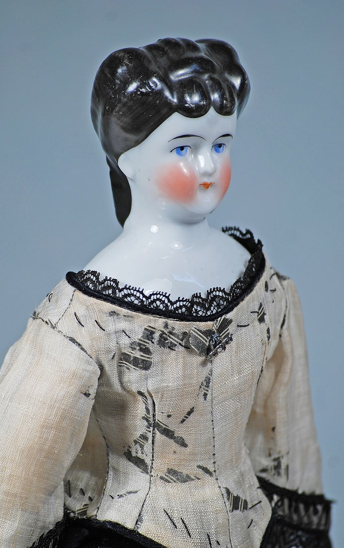 GERMAN PORCELAIN LADY WITH UNUSUAL COIFFURE: 14”. Porcelain shoulderhead, sculpted black hair with curls draped across the forehead and roll of widely-flared curls on each side which are pulled to the back into an arrangement of braids and