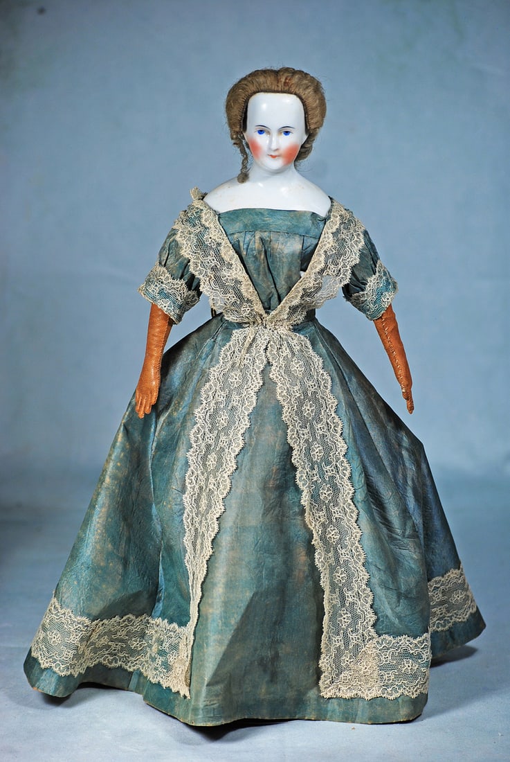 ELEGANT GERMAN PORCELAIN LADY WITH ORIGINAL WIG AND GOWN: 13 ½”. Porcelain bald head, solid-dome shoulderhead with original light brown mohair wig, heart-shaped face, painted features, blue eyes, red and black eyeliner, arched and tapered brows, c