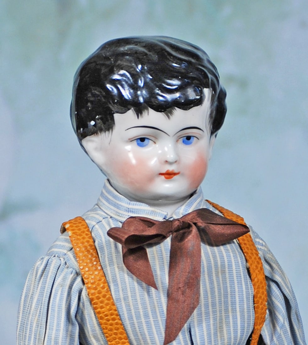 GERMAN PORCELAIN DOLL AS YOUNG GENTLEMAN: 16”. Porcelain shoulderhead, sculpted black hair with side-swept waves and curls, detailed brush marks at forehead and sides, modeled ears, painted features, shaded blue eyes, red and black eyel