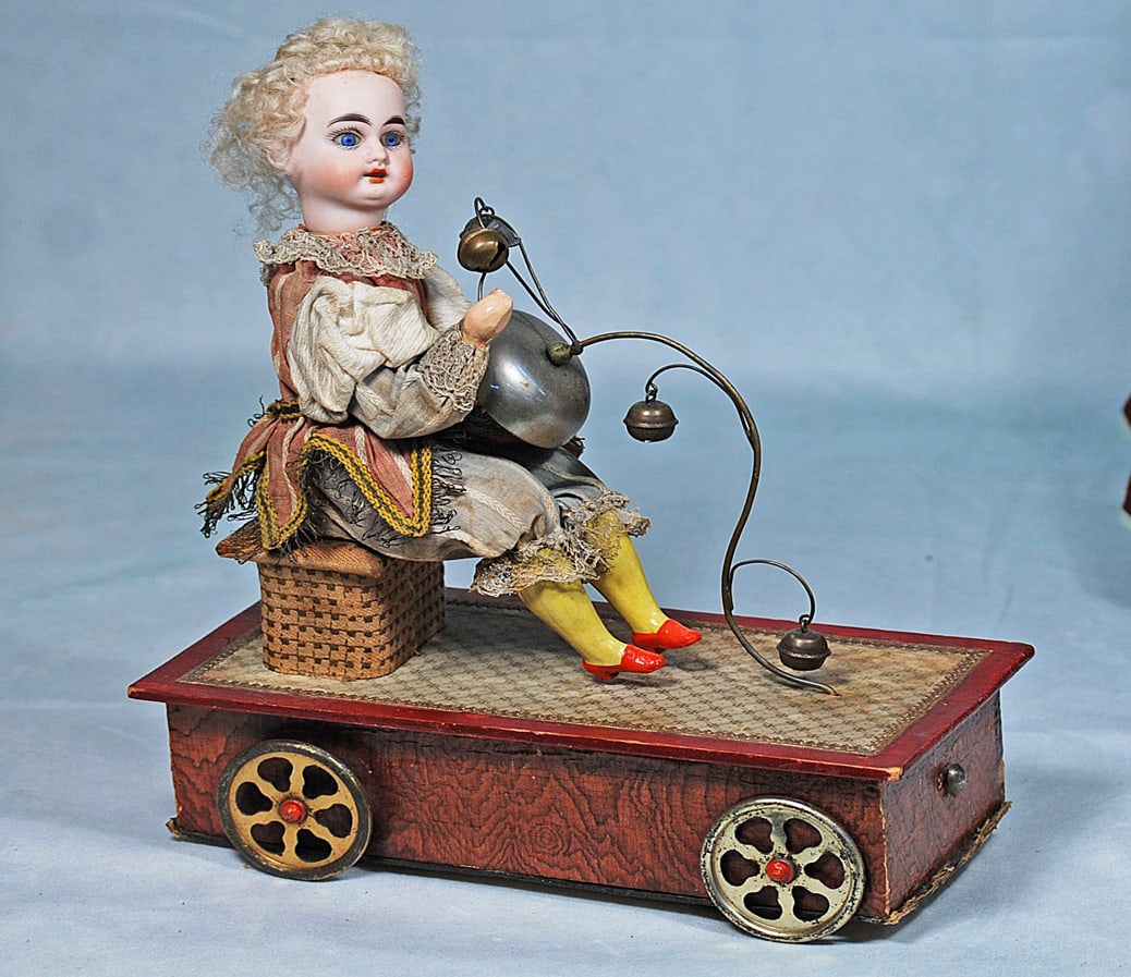 GERMAN BISQUE “JESTER WITH BELLS” MUSICAL MECHANICAL PULL TOY ATTRIBUTED TO ZINNER AND: 11” H. Posed upon a wooden platform with lithographed paper cover is a bisque head doll with inset blue glass eyes, painted lashes, feathered brows, open mouth with upper teeth, original blonde 