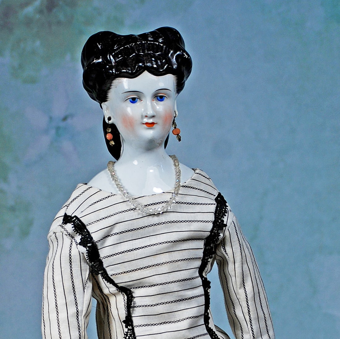 GERMAN PORCELAIN LADY WITH RARE COIFFURE, LARGE EYES AND PIERCED EARS: 22”. Marks: V. Porcelain shoulderhead, oval face with painted large blue eyes, red and black eyeliner, tapered brows, closed mouth, pierced ears, sculpted black hair arranged in flowing waves an