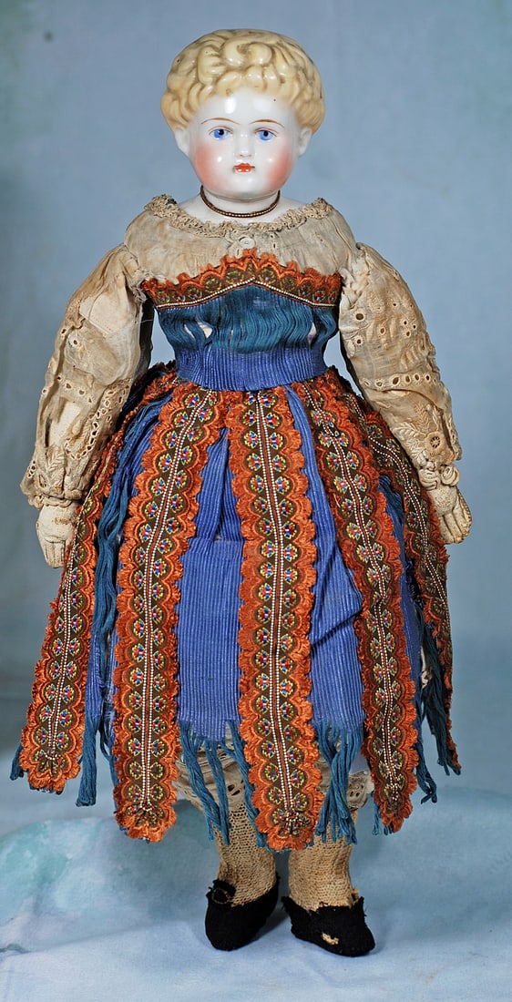 GERMAN PORCELAIN DOLL IN FOLKLORE COSTUME: 16”. Porcelain shoulderhead with sculpted short blonde hair arranged in abundant curls all over her head, sculpted ears, painted features, blue eye, black and red eyeliner, plump cheeks, closed 