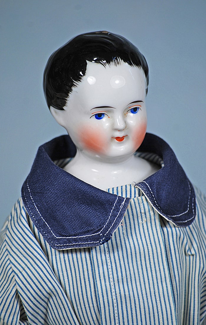 GERMAN PORCELAIN DOLL AS YOUNG GENTLEMAN: 15”. Porcelain shoulderhead with sculpted black hair in boyish manner with stippling detail around the face, fully modeled ears, painted features, deep blue eyes, red and black eyeliner tapered