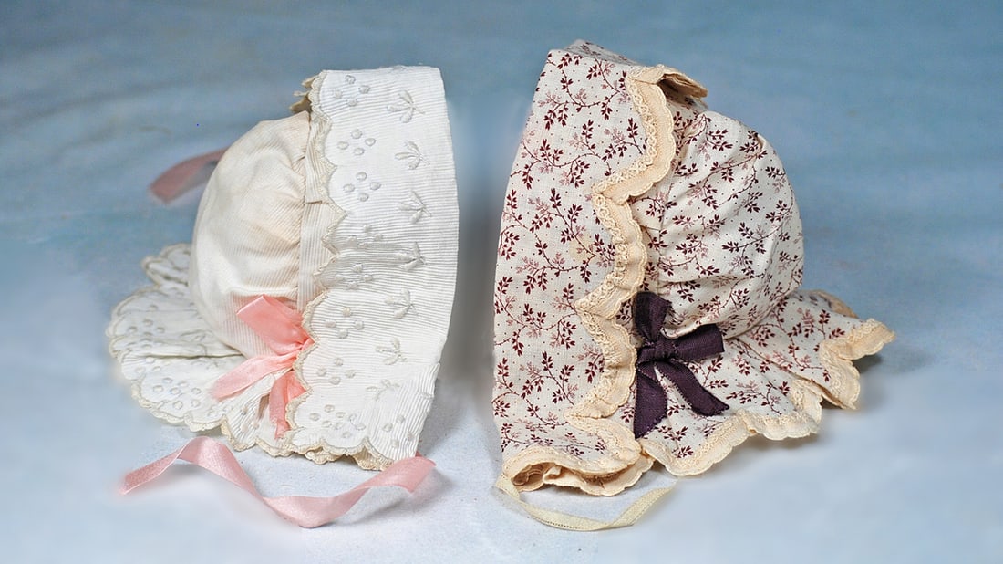 TWO DOLL BONNETS: Each is hooded bonnet with bavolet and ribbon ties. White cotton pique bonnet with dainty scalloped edging all around and embroidery detail, pink silk bows and ties; white and maroon cotton print