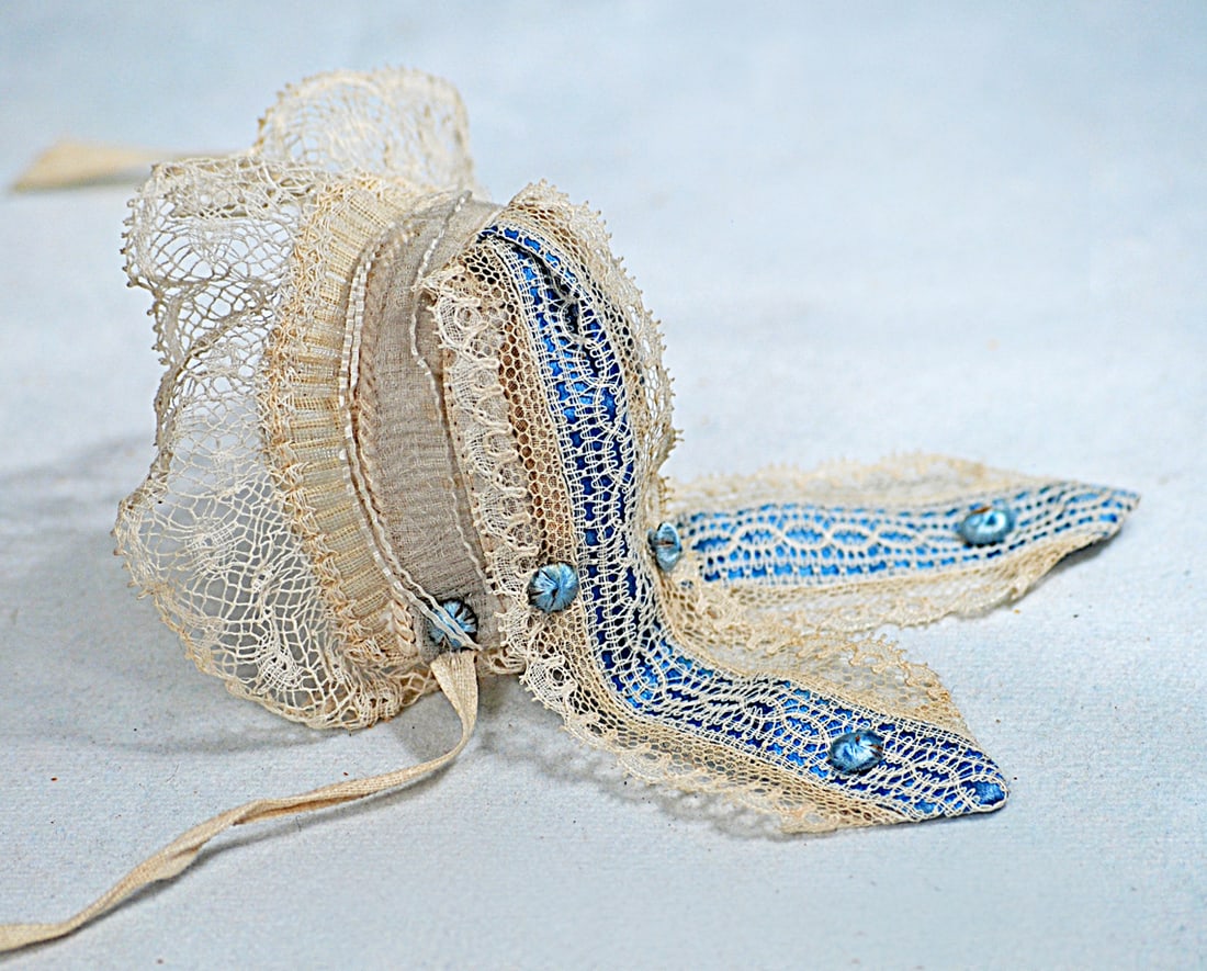 FINE ANTIQUE ORGANDY DOLL BONNET: White organdy made up in a dainty bonnet trimmed with val lace and tiny arranged blue silk balls, blue ribbons edged with lace forming elegant pendant barbes which fall long and gracefully over the ba