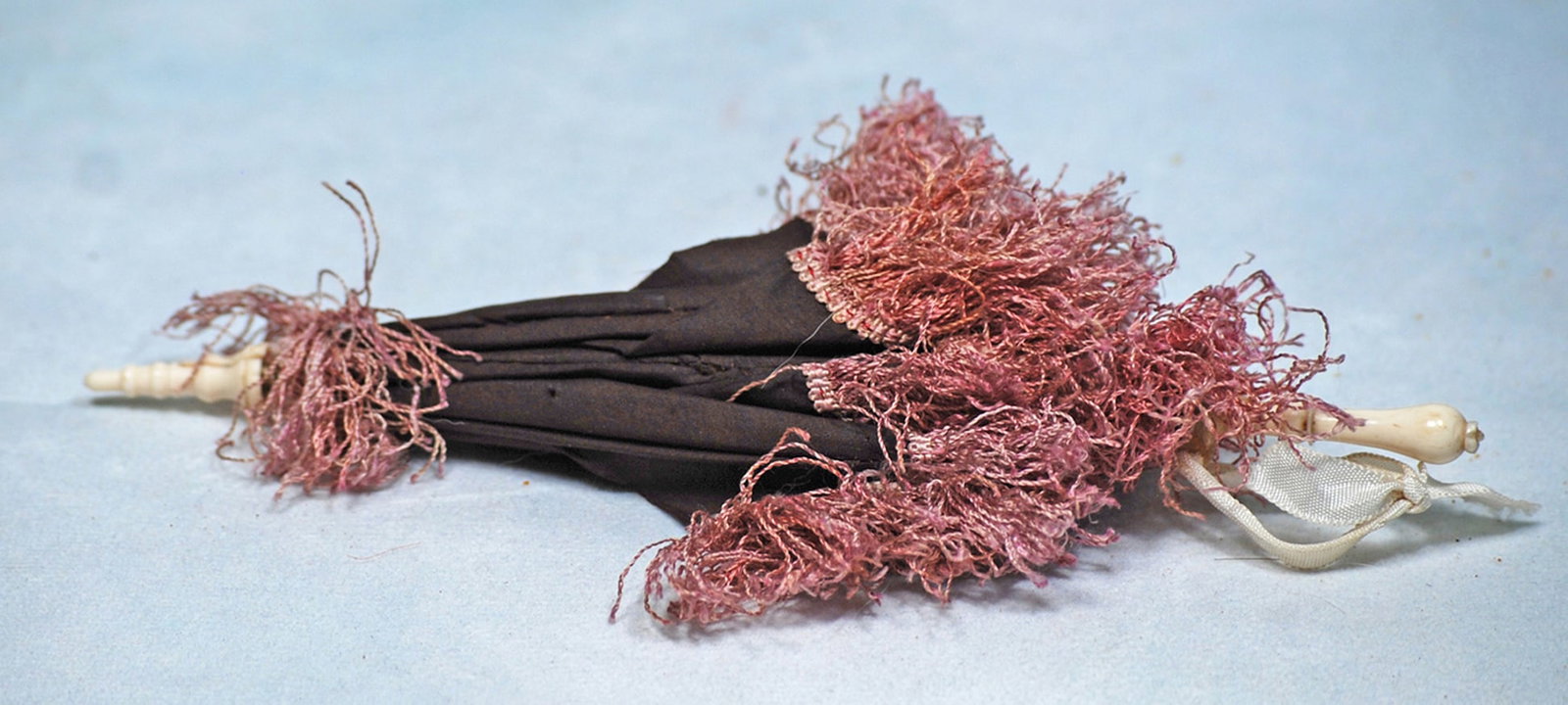 ANTIQUE DOLLS SILK PARASOL: 8”. Dark maroon silk parasol decorated with thick lavender crimped fringe, turned bone handle and tip, metal fitting.