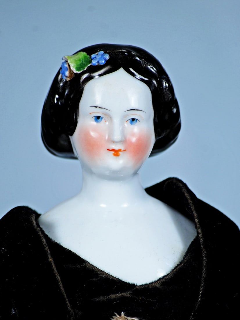 GERMAN PORCELAIN LADY WITH FLORAL DECORATED HAIR: 14”. Porcelain shoulderhead of lady with sculpted black hair adorned with applied blue flowers, hair center-parted and waved along the sides and drawn to the back, arranged in a coiled chignon, 