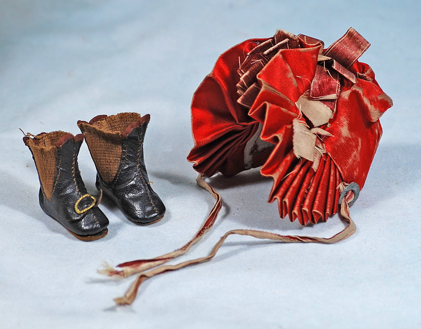 ANTIQUE DOLL BONNET AND BOOTS: Red silk bonnet with pleated 1 ½” deep graduated brim, pleated back garnished with clusters of maroon silk bows, buckram lining, silk ribbon ties, for about 8” head circumference; pai