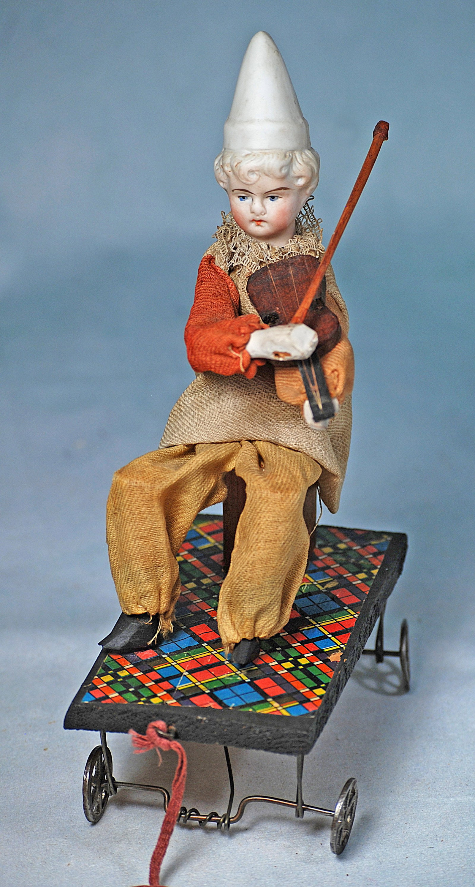 GERMAN PULL TOY CLOWN WITH FIDDLE: 9” x 5 ½”. Bisque head clown with sculpted pointed hat over blonde curls, painted features, wooden torso, hands and feet, posed on wooden wagon, holds carved wooden fiddle and bow; wh