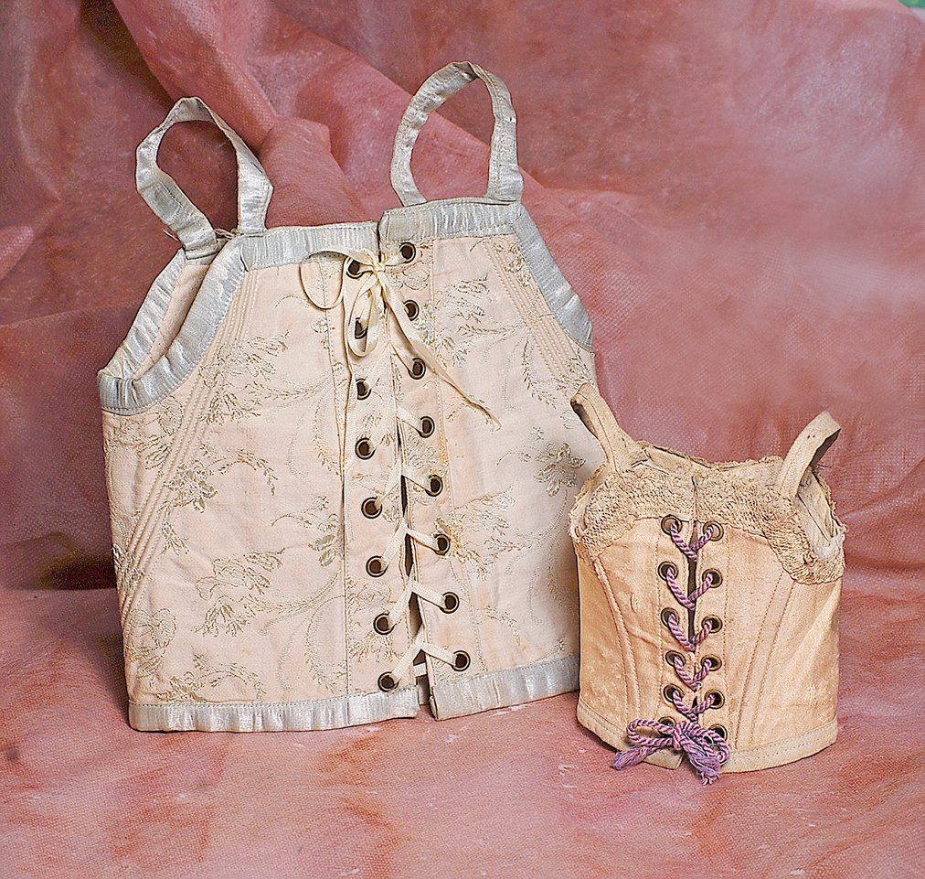 TWO ANTIQUE FRENCH BEBE STAYS. (1) Cream silk Jumeau: TWO ANTIQUE FRENCH BEBE STAYS. (1) Cream silk Jumeau bebe stay with delicate pale blue floral embroidery, blue silk edging and straps, sixteen brass-edged lacing grommets, 14” waist circumference, 9