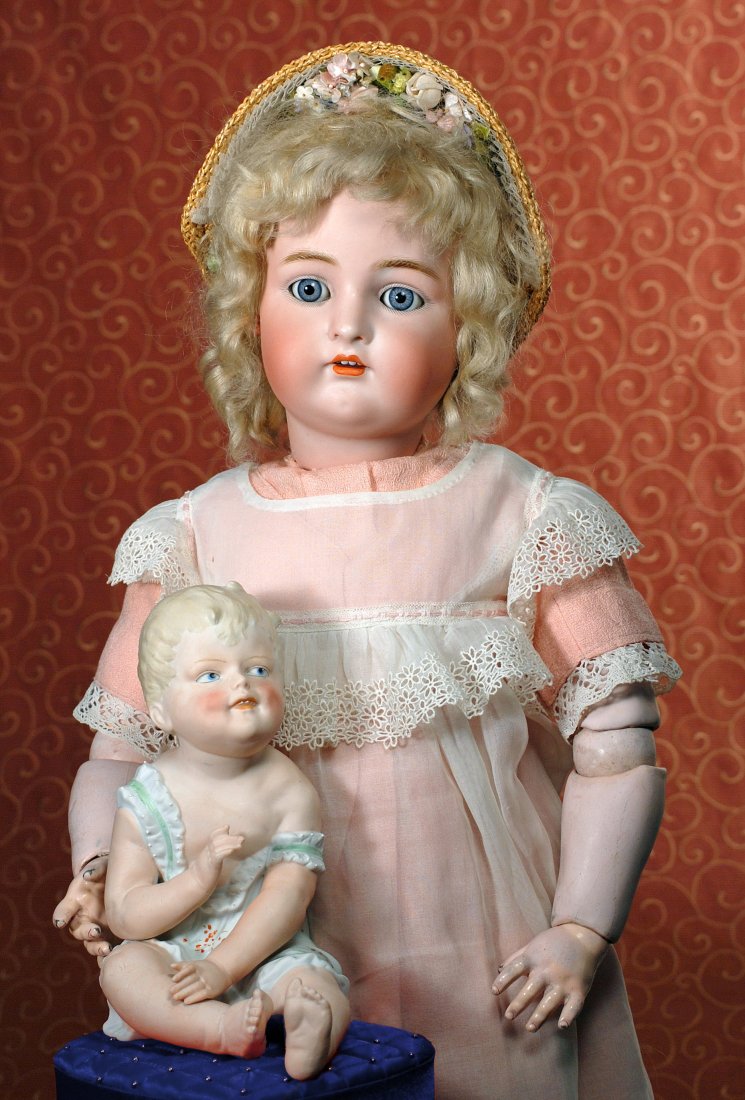GERMAN BISQUE PIANO BABY: GERMAN BISQUE PIANO BABY. 8 ½”. Seated bisque baby has sculpted and swept forward blonde hair, painted blue eyes with gray eyeliner all-around, lightly feathered brows, deep dimples, open/closed mo