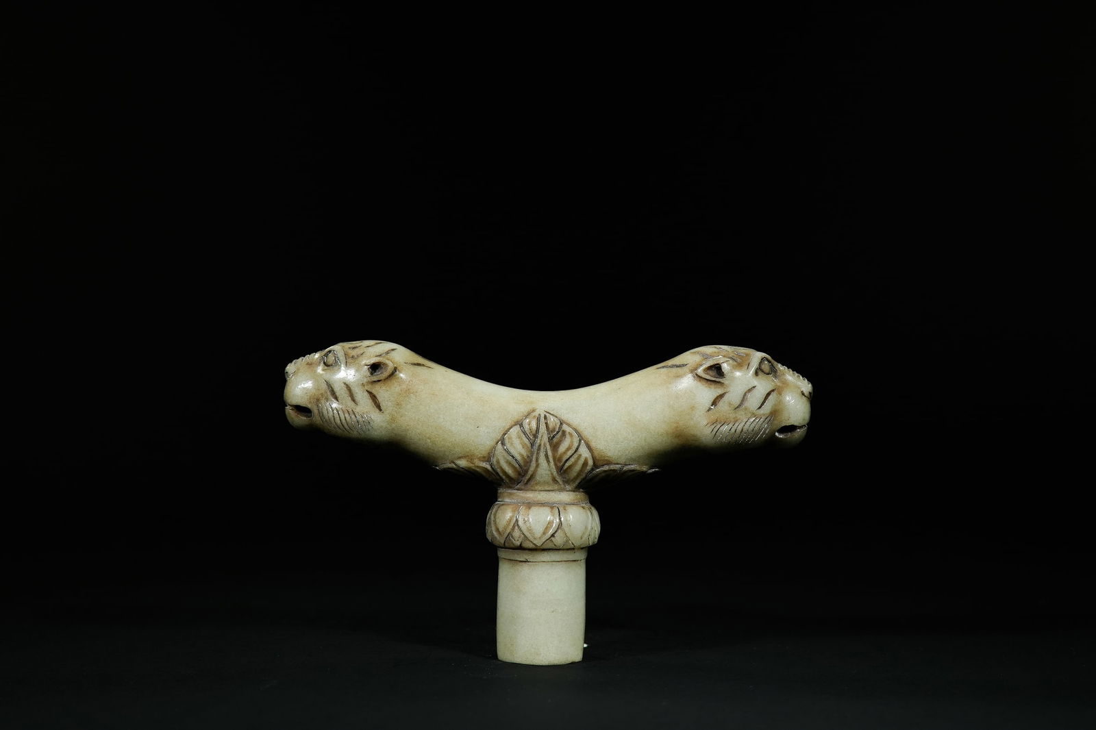 A Mughal Jade Dagger Hilt with Twin Feline Heads: A Mughal jade dagger hilt carved in a pale, creamy green stone, shaped with an elegant outward curve. The upper section features two opposing feline heads, each rendered with defined eyes, ridged muzz