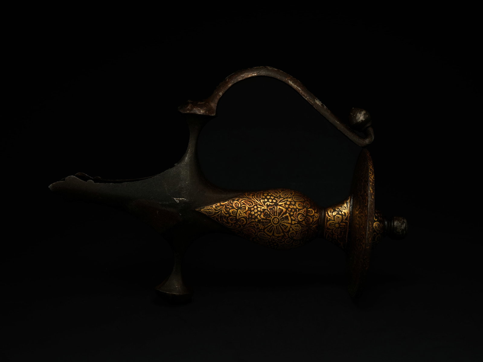 A Mughal Steel and Gold-Inlaid Tulwar Hilt, 19thÃ¢â‚¬â€œ20th Century.: The hilt is formed entirely of darkened steel and richly decorated with gold inlay, arranged in dense floral and foliate scrollwork. The grip curves elegantly into a zoomorphic pommel, the surface art
