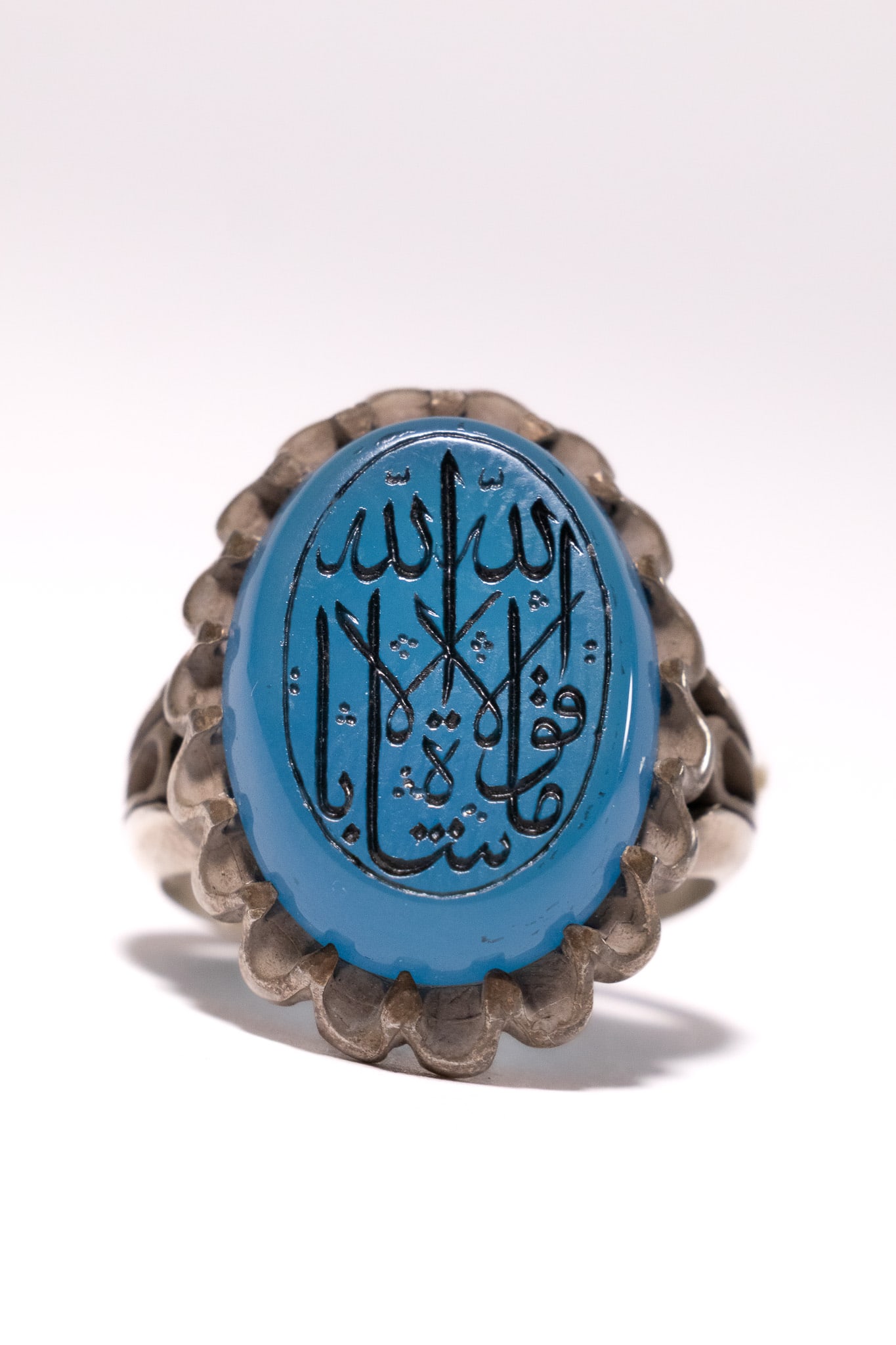 AN ISLAMIC SILVER AND BLUE AGATE SIGNET RING WITH ARABIC CALLIGRAPHY.: An Islamic signet or amulet ring featuring a silver metal setting and a prominent oval blue cabochon stone. The stone is decorated with intaglio Arabic calligraphy, featuring the word Allah at the top