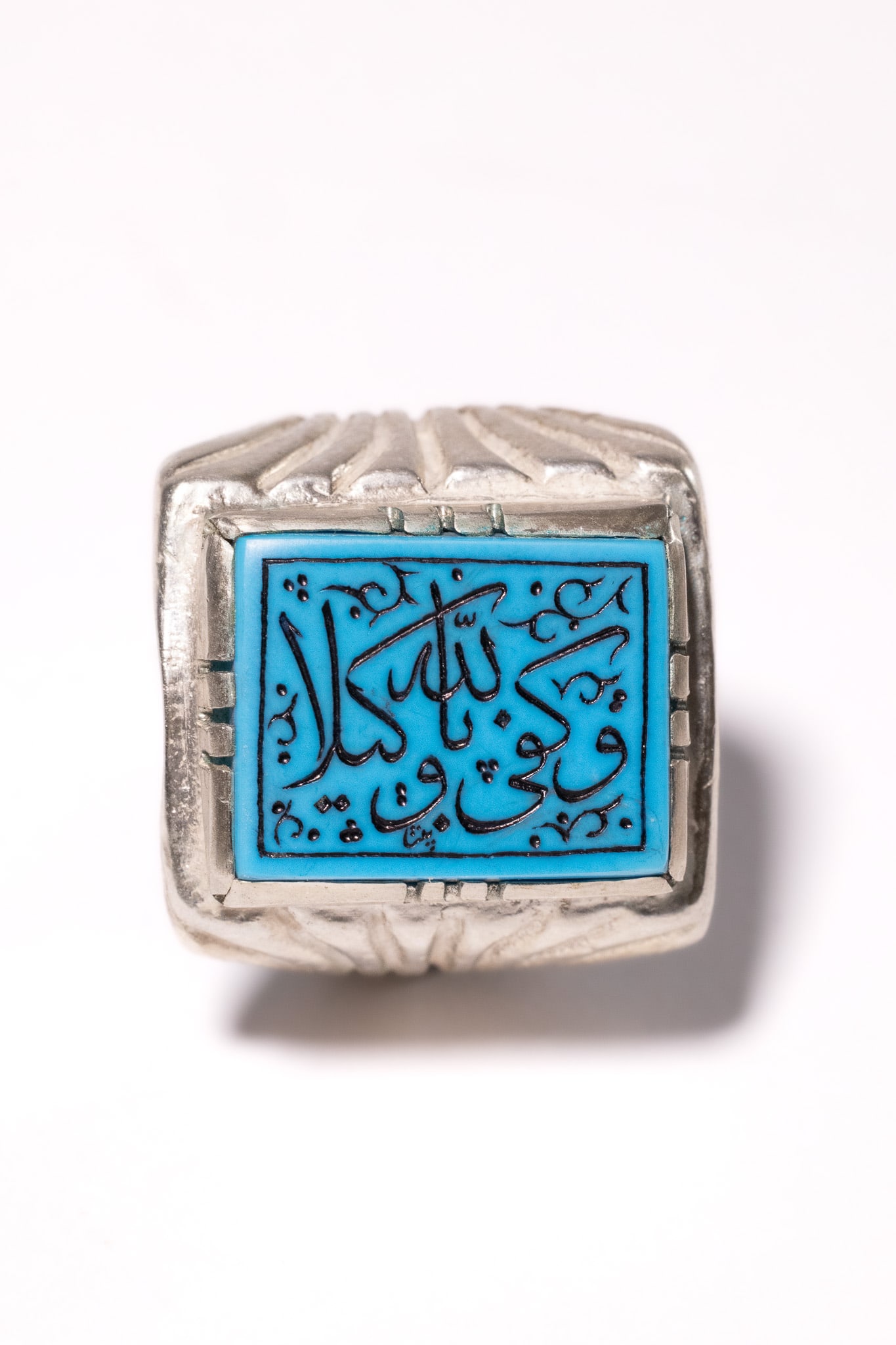 AN ISLAMIC SILVER SIGNET RING WITH ENGRAVED TURQUOISE-COLOURED AGATE.: The ring features a central rectangular tablet of turquoise-coloured agate. The stone is flat-topped and bears an incised Arabic inscription filled with black pigment. The inscription is the Quranic p