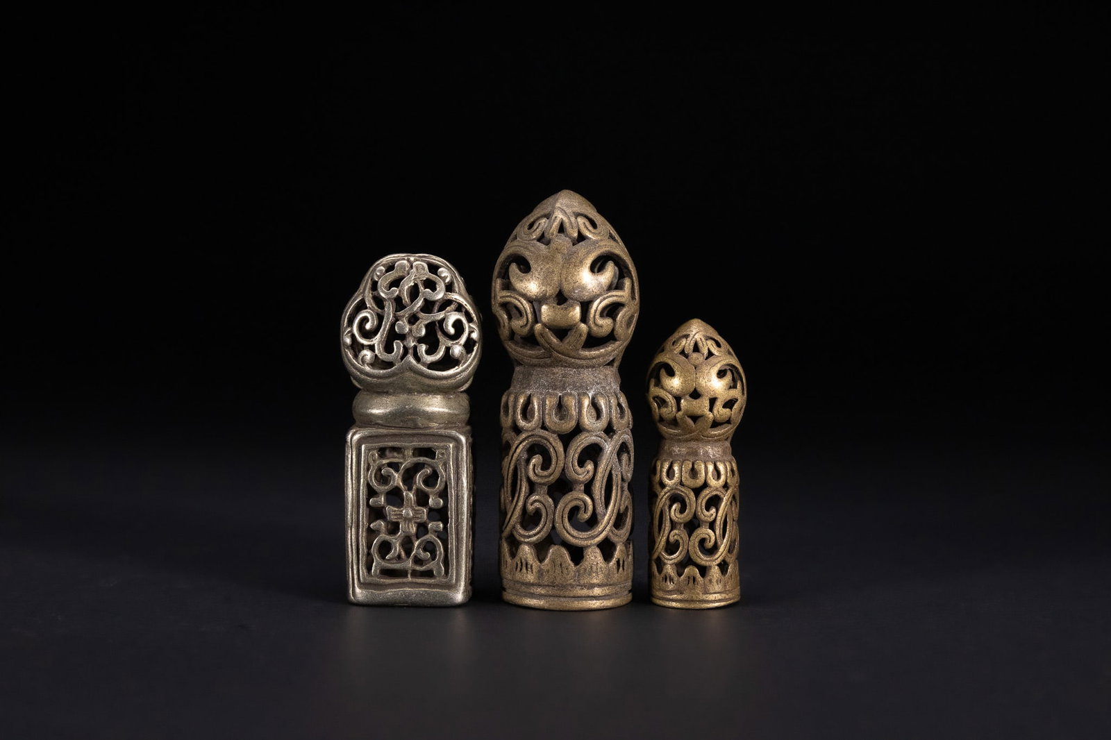 A GROUP OF THREE TIBETAN BUDDHIST CAST METAL OPENWORK CHOP SEALS.: This lot comprises three cast metal decorative seals of Himalayan origin, specifically attributed to Tibetan Buddhist culture. The set includes one silver-toned seal with a square base and two brass-t