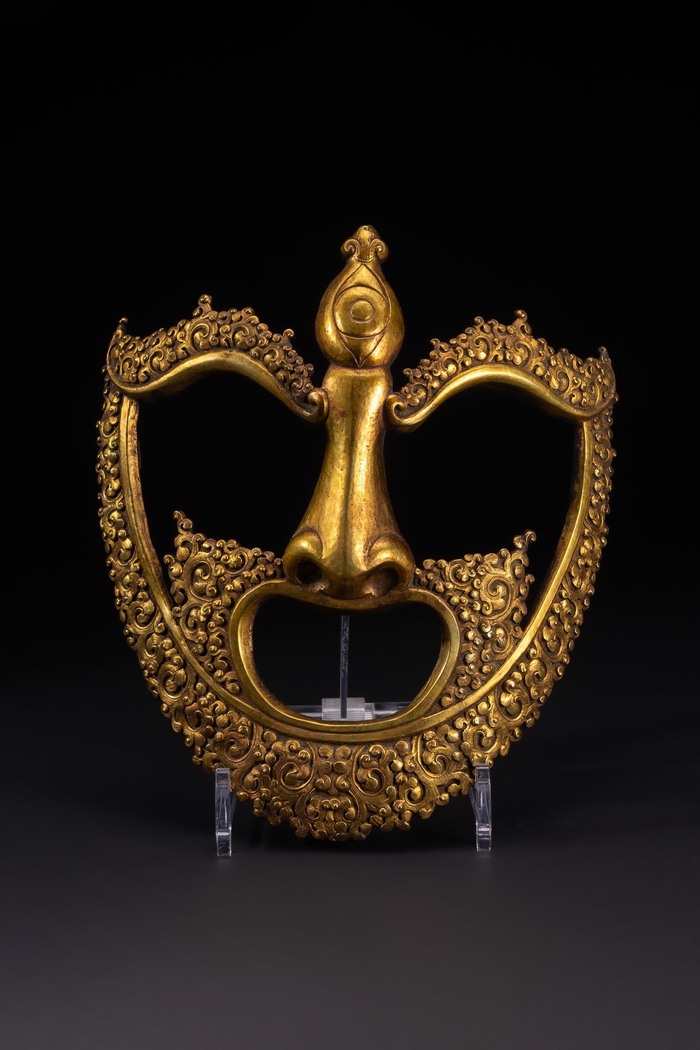 A TIBETAN GILT BRONZE RITUAL MASK OR KIRTIMUKHA ELEMENT.: A cast copper alloy ritual mask or architectural element, likely representing a Kirtimukha or Face of Glory. The object is finished with a heavy layer of gilding to the front surface. The iconography