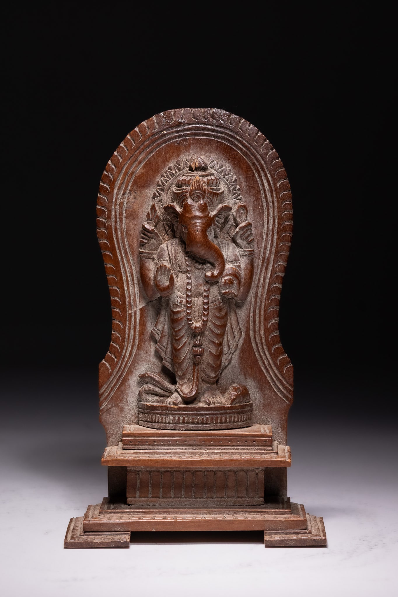 A SOUTH INDIAN CARVED HARDWOOD RELIEF SCULPTURE OF GANESHA, 20TH CENTURY.: This carved hardwood relief sculpture depicts the Hindu deity Ganesha in a standing sthanaka posture. The figure is positioned upon a circular lotus pedestal mounted on a tiered rectangular architectu