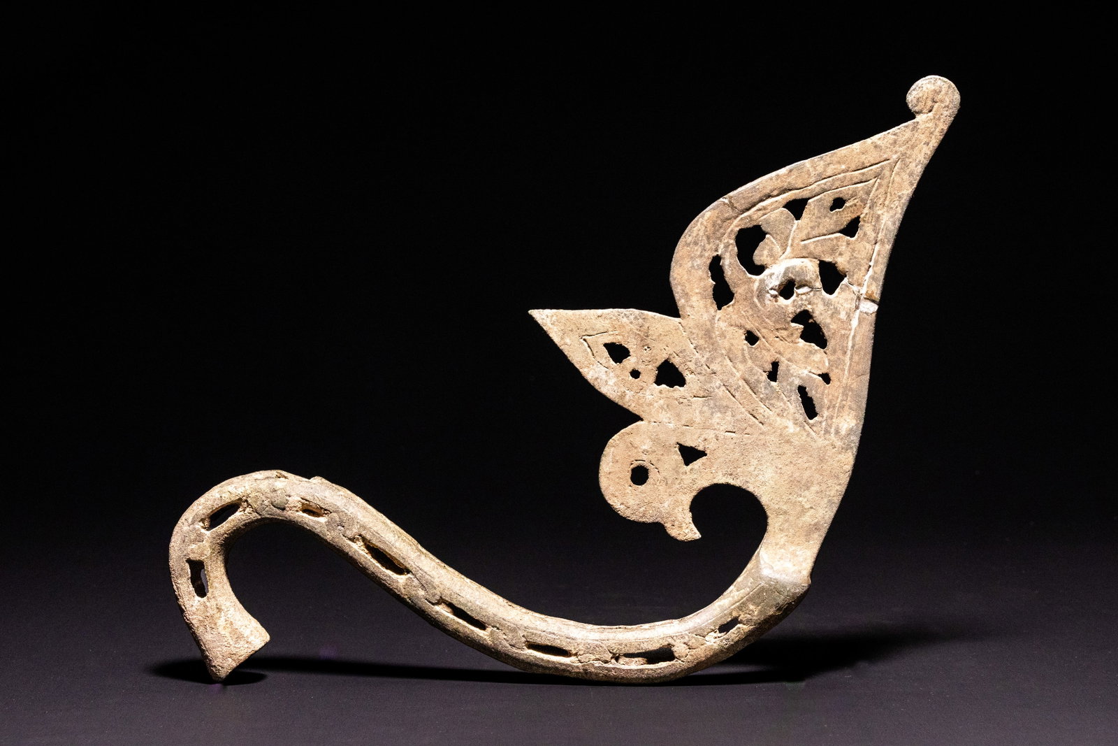 AN ISLAMIC BRONZE OPENWORK FOLIATE FITTING OF AN INCENSE BURNER, 12TH CENTURY.: A bronze fitting featuring a long, gracefully curved square-sectioned arm. The arm is decorated with a series of recessed rectangular piercings along its outer edge, terminating in a hook at the lower