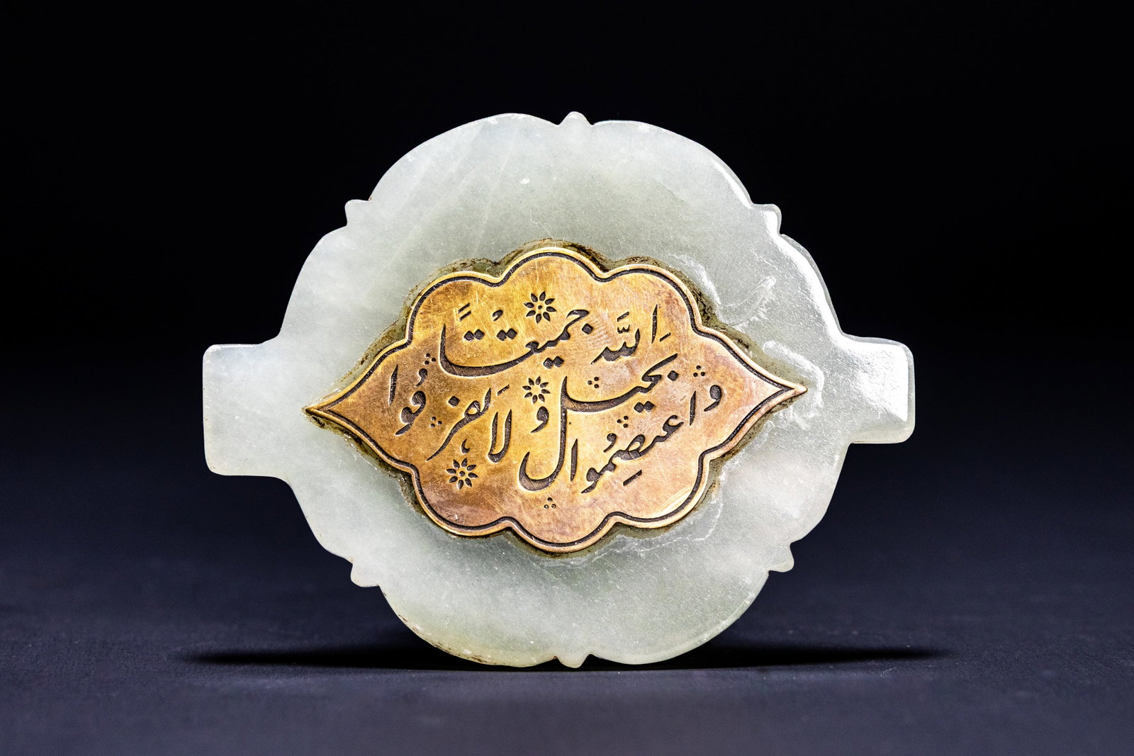 AN INDO-PERSIAN INSCRIBED PALE GREEN JADE AND COPPER INLAID PLAQUE, 19TH CENTURY OR LATER.: An inscribed decorative plaque carved from semi-translucent pale green nephrite jade featuring a central flush-mounted copper alloy inlay. The jade body is shaped with a lobed foliate outline in the c
