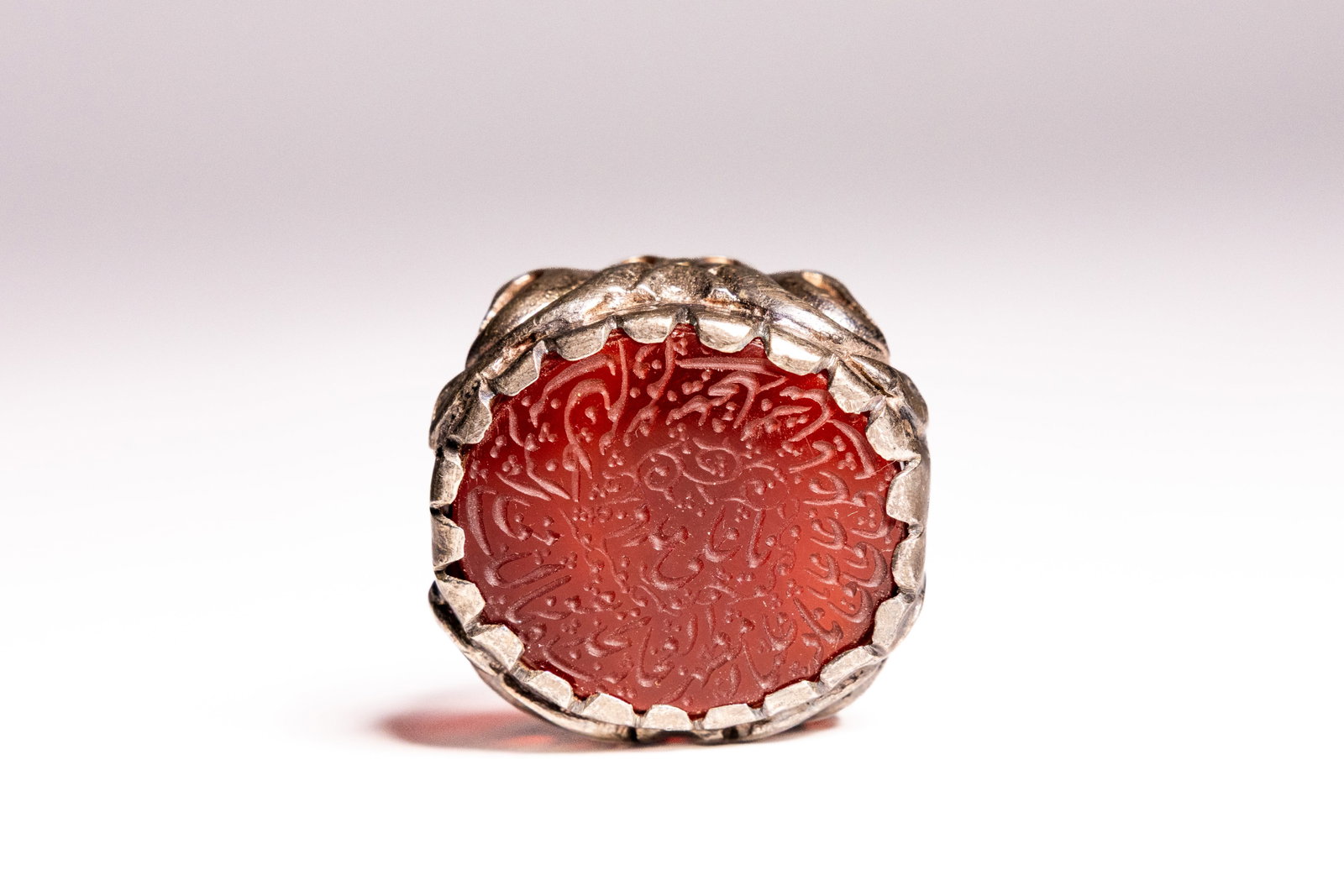 AN ISLAMIC SILVER AND CARNELIAN INTAGLIO CALLIGRAPHIC SEAL RING.: An Islamic seal ring featuring a central oval carnelian gemstone, often referred to as aqeeq. The translucent reddish orange stone is intricately hand engraved with dense Arabic calligraphy in a circu