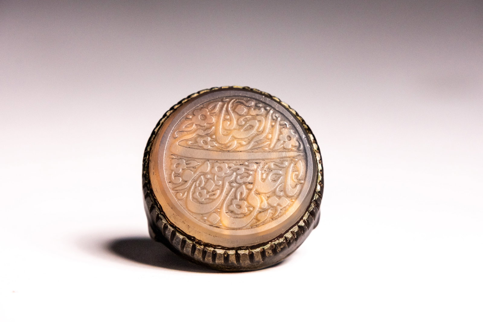 AN ISLAMIC SILVER AND AGATE INTAGLIO SEAL RING.: An Islamic intaglio seal ring featuring a central oval translucent agate stone showing natural honey-coloured banding. The stone is deeply incised with calligraphic script arranged in two horizontal r